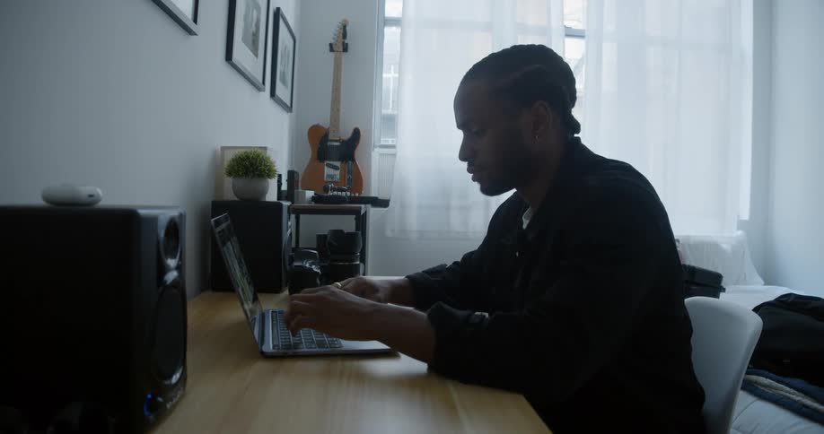 A man is sitting at a desk and working on a laptop at home in his bedroom during the daytime