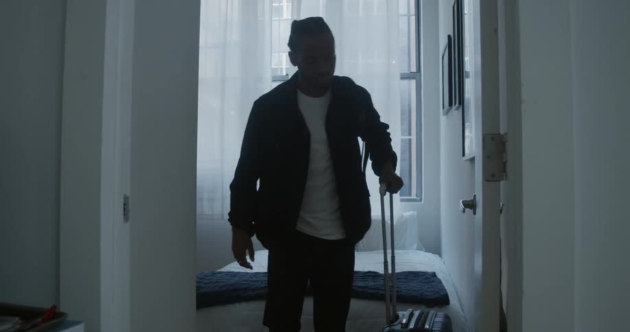 Man picks up backpack and rolls suitcase out of bedroom