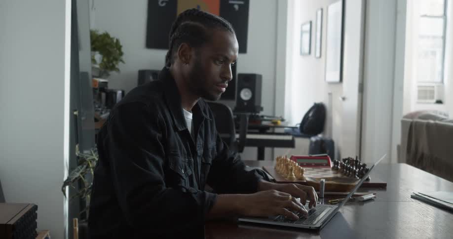 Man with braids and black jacket sits at table working on laptop next to chess