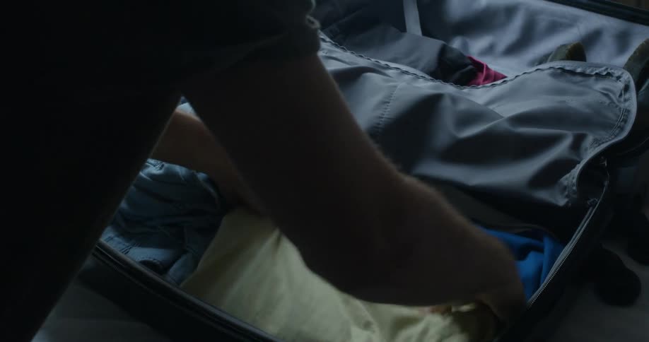 Man folding clothes inside luggage over a bed in a room