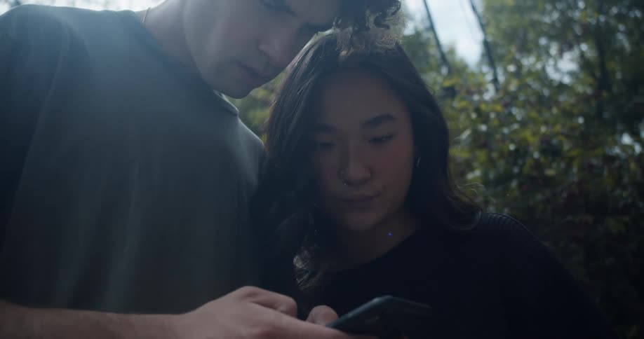 Man with curly hair and green t shirt and woman with straight hair and black sweater use smartphone in the forest