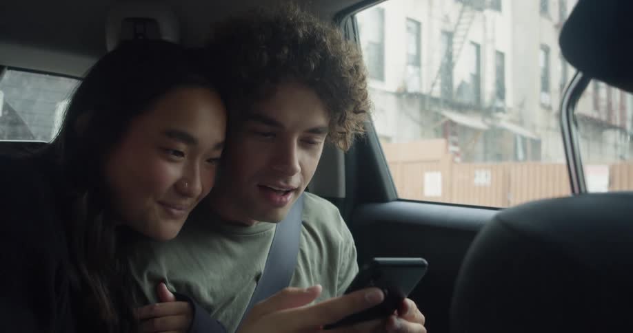 Man with curly hair and green t shirt and woman with straight hair and black sweater sit in the back seat of the car and take selfie with smartphone