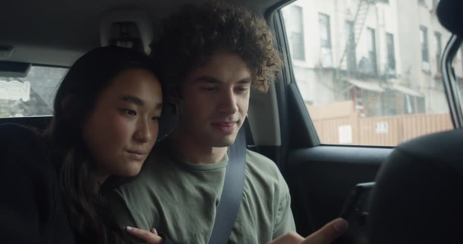 Man with curly hair and green t shirt and woman with straight hair and black sweater takes selfie with a smartphone in car moving in the city street
