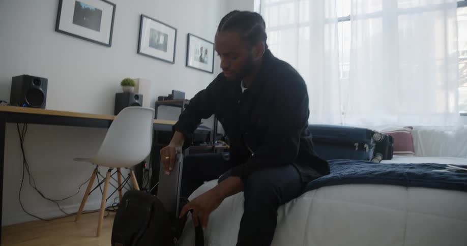 A man sitting on a bed packs his laptop into his backpack