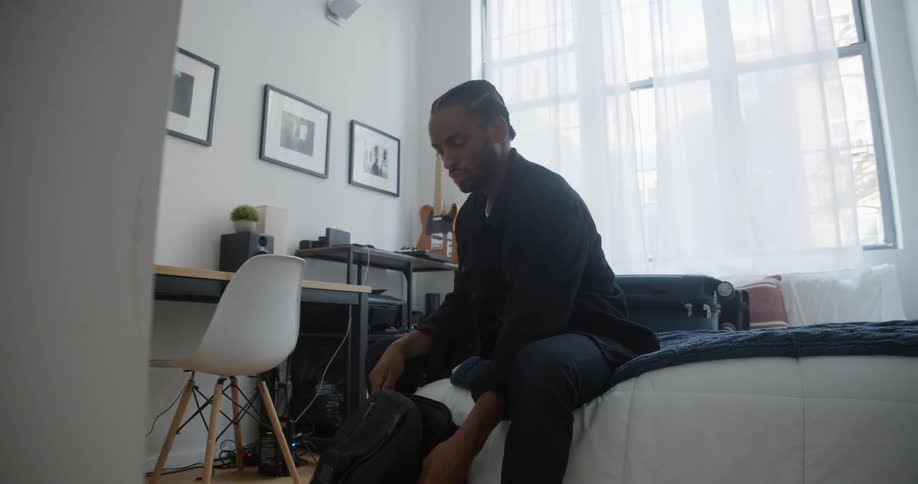 Man with braids black jacket and pants sitting on bed zips up backpack and gets up to leave in room