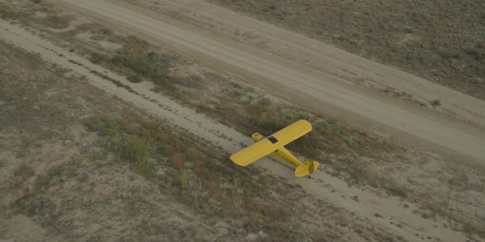 A yellow plane flies over a scrubby runway