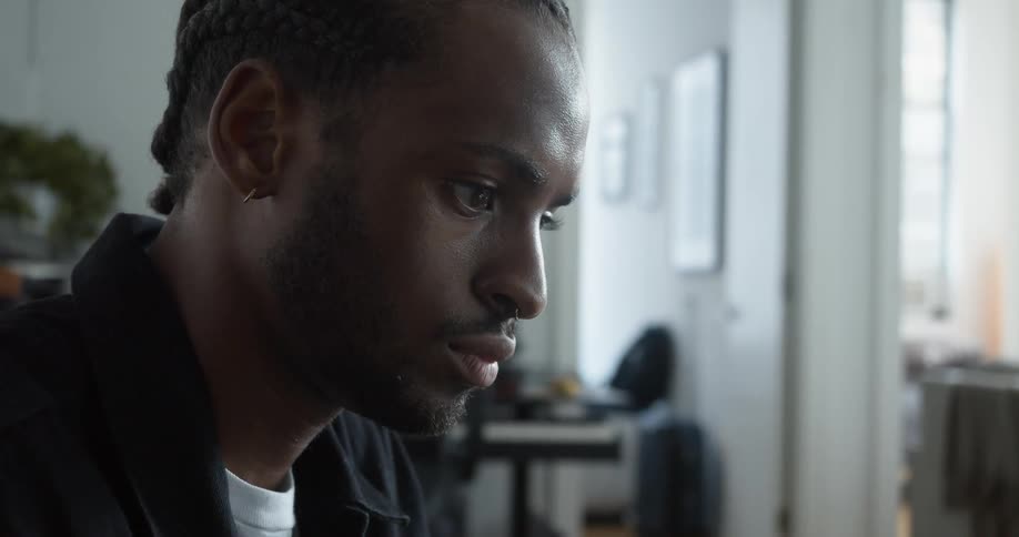 Man with braids earring beard and black jacket sits at desk and uses laptop in living room at home