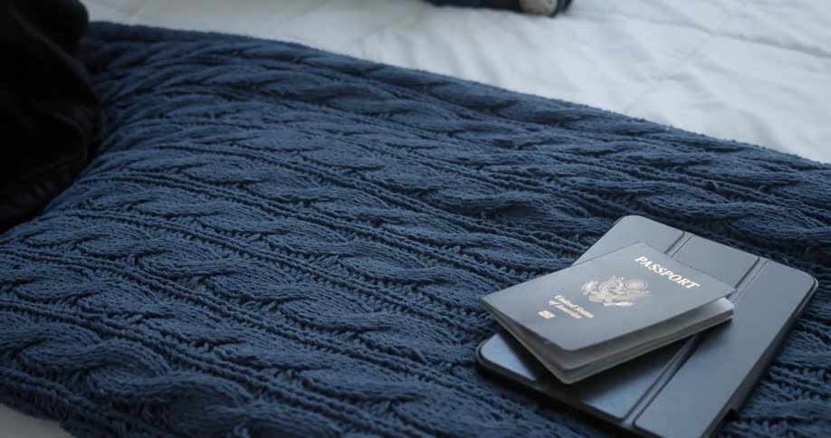 A man sits on his bed and packs away his laptop tablet and passport