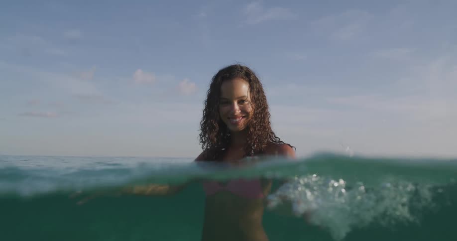 Woman with curly hair and pink bikini standing in sea splashes water smiling