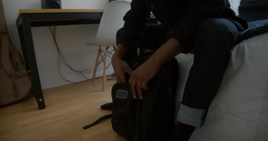 A man sitting on a bed packs his laptop in a backpack then flips through his passport before packing it