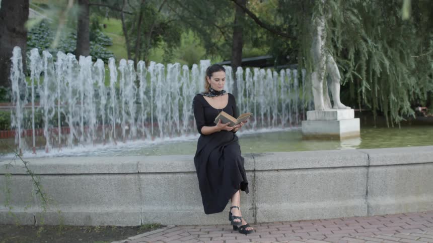 Woman wearing black dress sits on edge of fountain reading hardback book