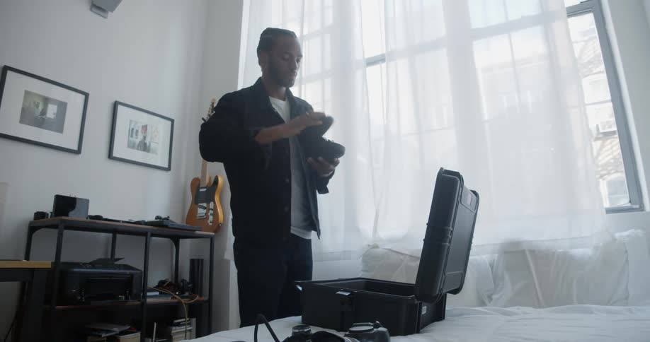 Male photographer packs camera into case sitting on bed near window