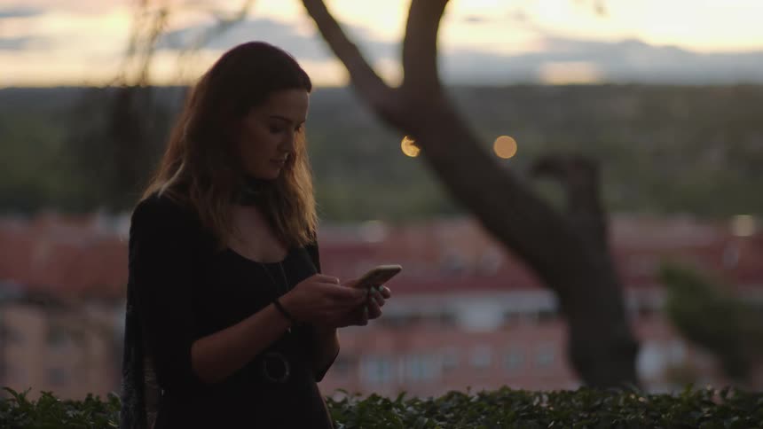 A woman is standing outside texting on her smartphone at dusk evening