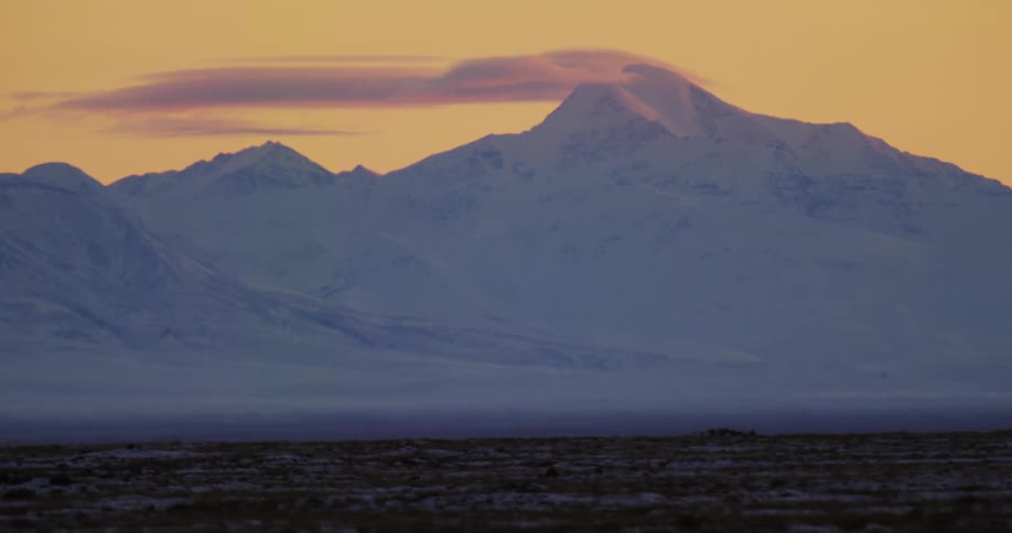 Tundra borders snow covered mountains that contrast with red sky
