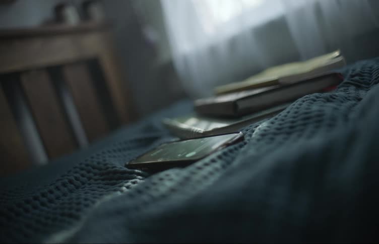 A bedroom during the day where books and a smartphone showing a video are laying on the bed