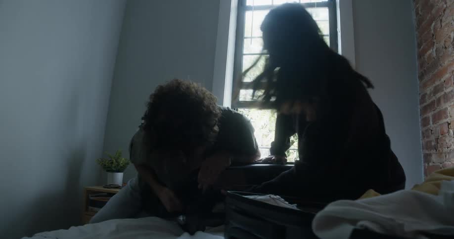 A couple laughs together as they try to force a stuffed suitcase closed