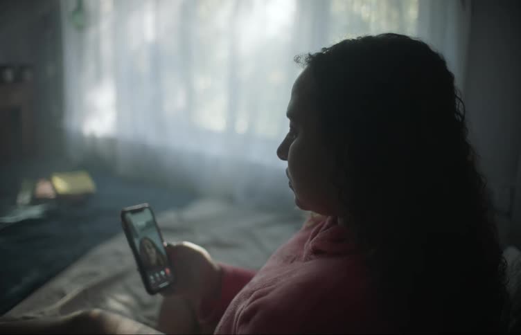 Woman sitting in shadowy room looks at herself in camera app before throwing phone onto bed
