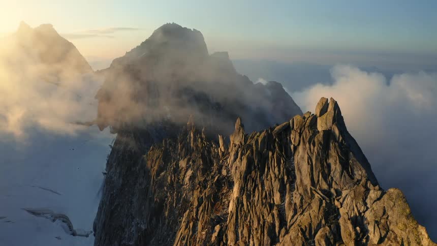 The sun shines brightly on a mountain ridge with peaks high in the clouds