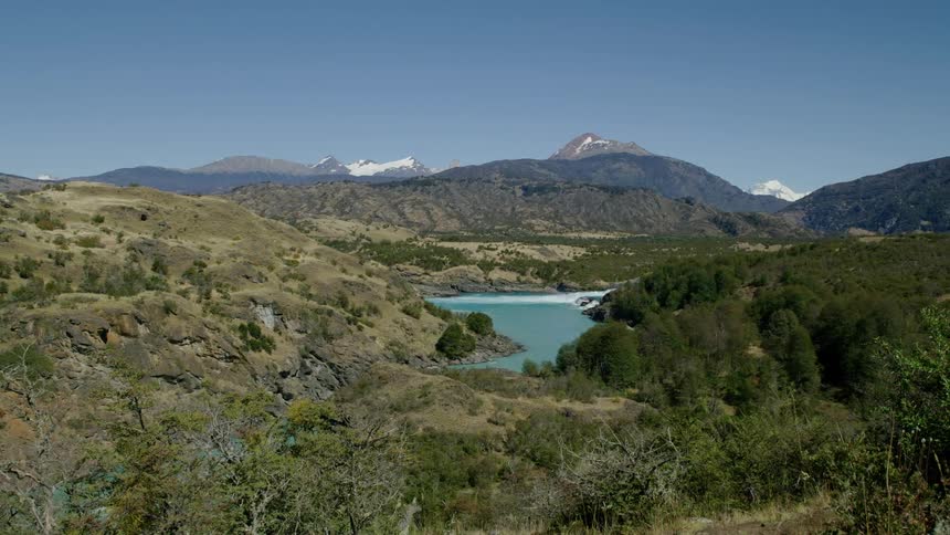 A river is flowing through a hilly landscape with mountains in the background on a clear and sunny day