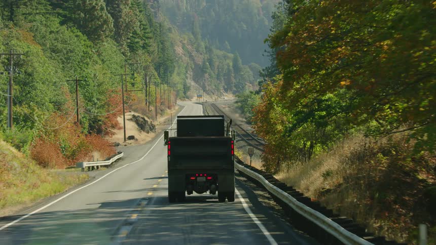 A dump truck drives down a two lane highway alongside train tracks in a classic Americana industry scene