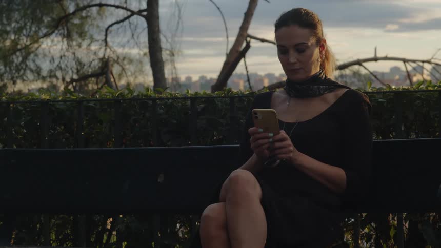 Woman sits on bench using mobile phone with city in background