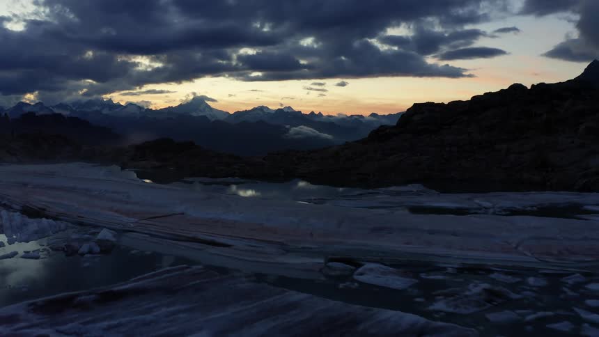 Ice covers water that reflects clouds with mountains on the horizon under a red tinted sky