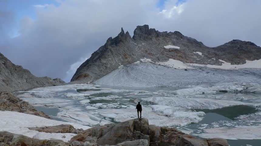 Aerial view of a man standing atop rocks in front of a frozen lake on a mountain peak