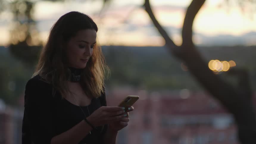A woman stands beneath a tree and looks at her cell phone as she texts