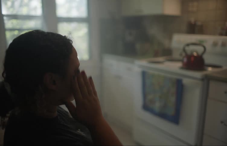 A woman sits and rubs her head then gets up to walk to the stove to take the kettle off the burner