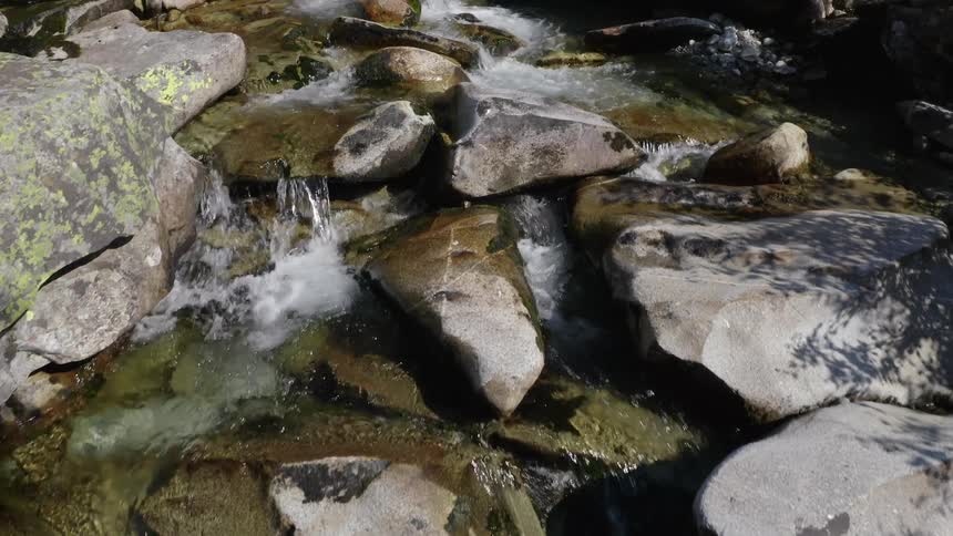Low altitude drone footage of a spring water stream flowing down a mountainside with a building in the distance during the day