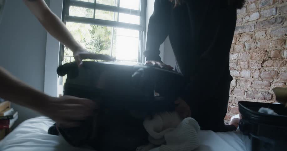 A man and woman laugh as they struggle to zip up an overstuffed suitcase