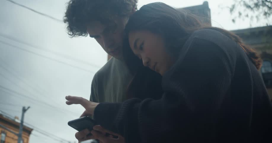 A young man and woman stand close together outside looking at a cell phone