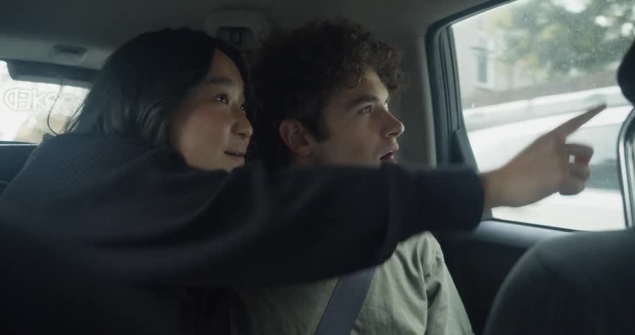 Man with curly hair and green t shirt and woman with straight hair and black sweater looks out the window of moving car in city streets