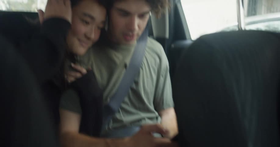 Woman with straight hair and black sweater holds man with curly hair and green t shirt using smartphone in car moving in the city street