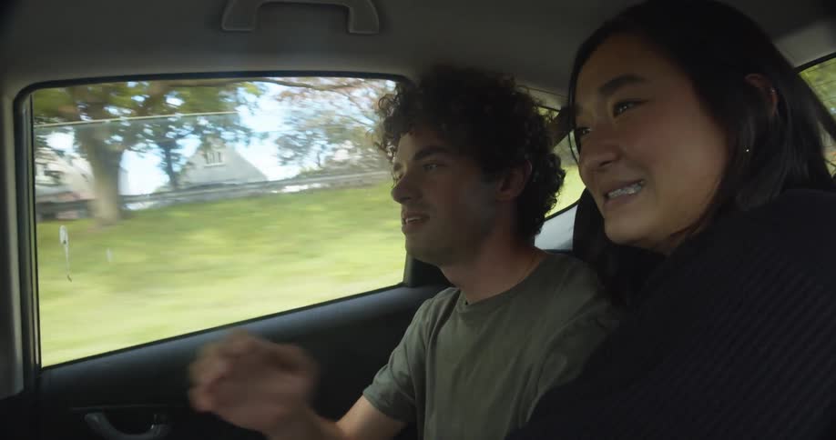 Man with curly hair and green t shirt and woman with straight hair and black sweater sit in the back seat of the car driving on highway