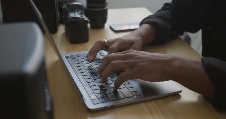 A man types on a laptop with his camera and phone on the desk