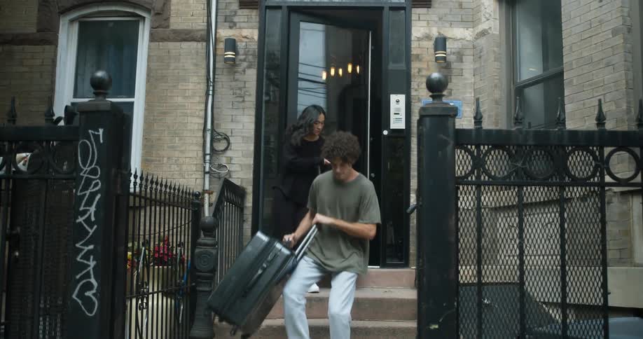 A man and woman are leaving an apartment building in a city with luggage in tow during the daytime
