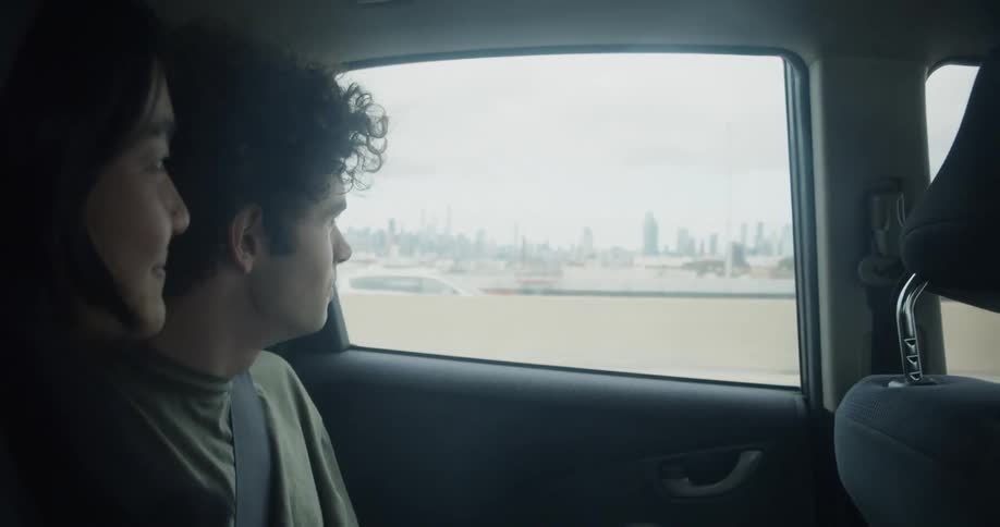 Happy man with curly hair and green t shirt and woman with straight hair and black sweater looks out the window of moving car on highway with city skyline on the horizon