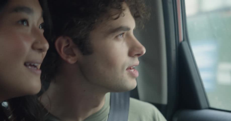Man with curly hair and green t shirt and woman with straight hair and black sweater looks out the window of moving car in city streets