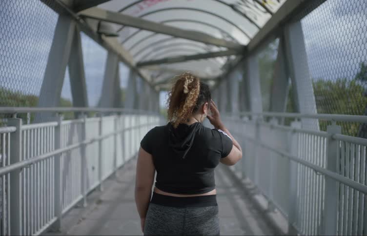 A woman walks across a pedestrian bridge and puts her hand to her head as she stops