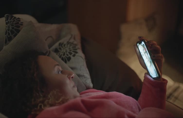 Woman with brown curly hair and pink bathrobe lies on bed making a video call on smartphone