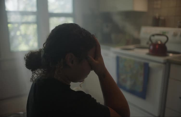 A woman stands in her kitchen as a tea kettle steams on the stove and rubs her forehead