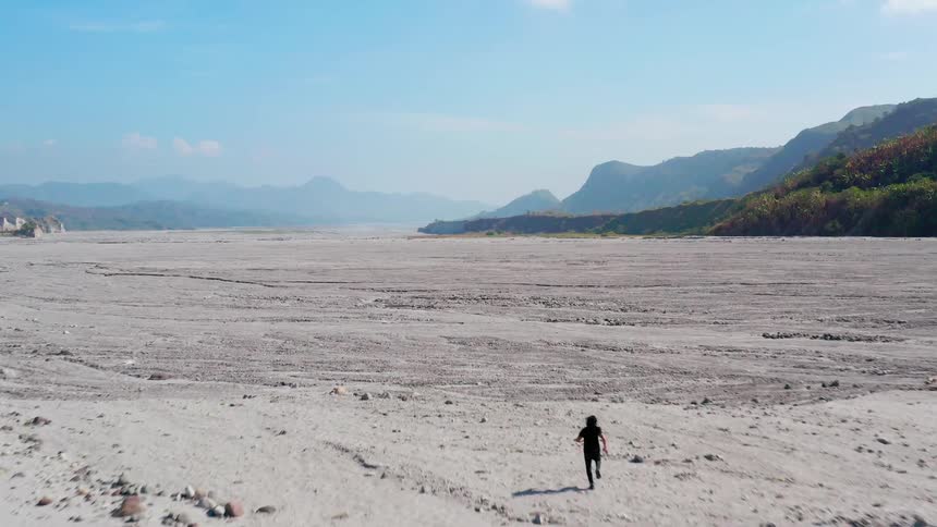 An aerial view of a man running in a large dried riverbed with a forest and mountains in the background
