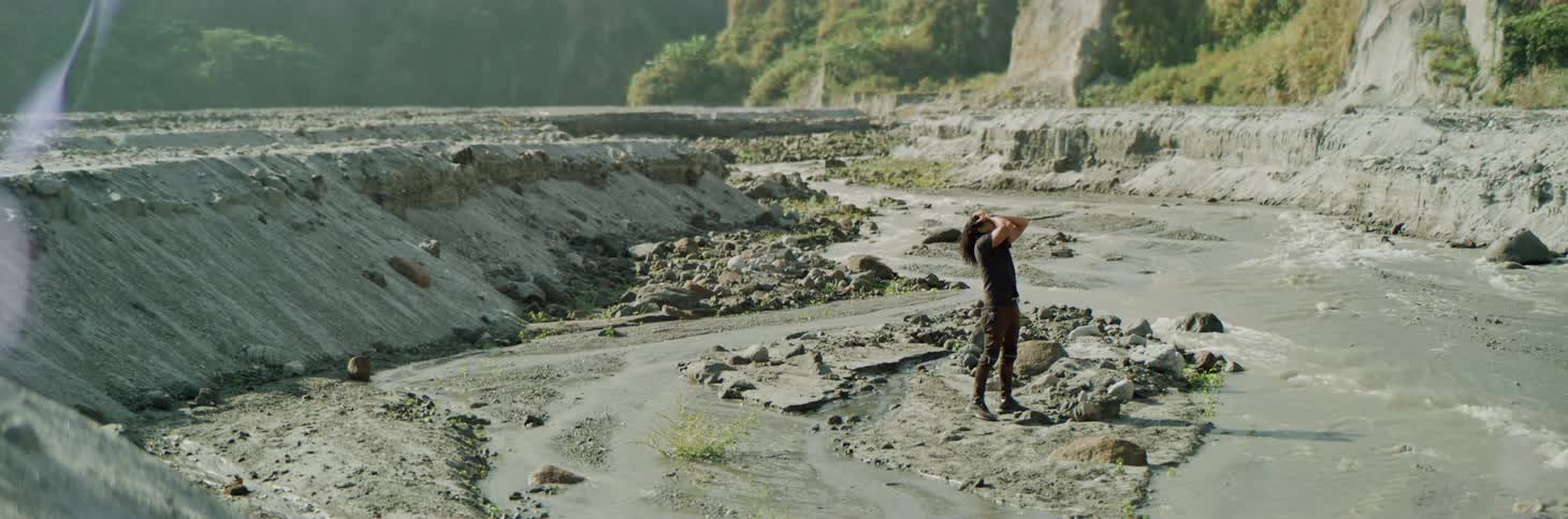 Man stands on dirt near flowing stream before bending over hands on knees