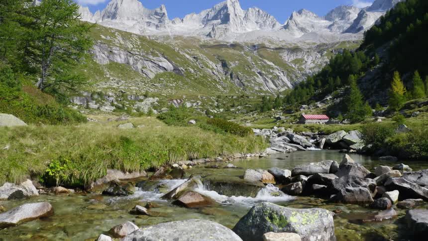 A low altitude view of a stream filled with rocks and boulders in a grass field with a house and mountains in the background
