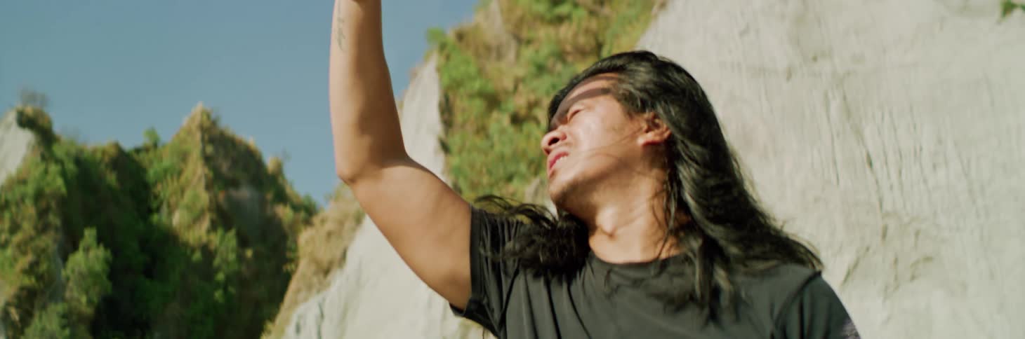 Man with long hair standing near mountain raises hand to shade his eyes