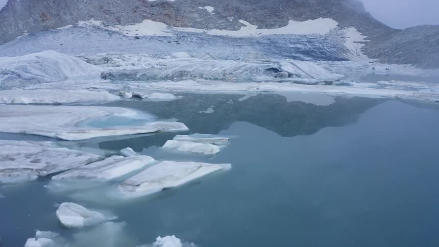 Ice covers ground and shore near water reflecting sky with mountains in background