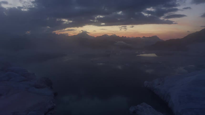 Fog covers ice floating on reflective water as mountains on horizon sit under a red sky