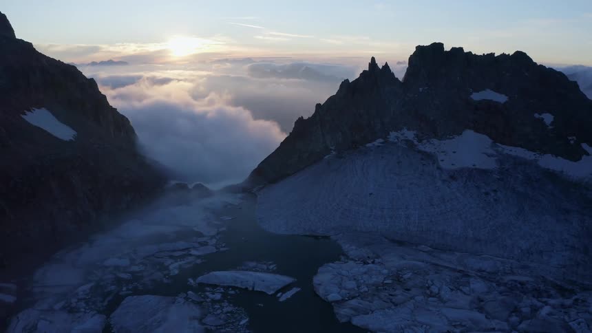 Snow covered mountains with peaks high in a cloudy sky and the sun shining over the clouds in the distance