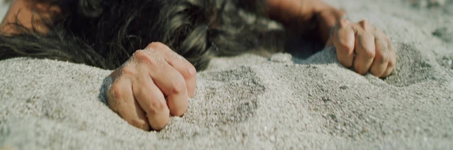 A person lying face down in the sand shakes and clenches their fists around handfuls of sand